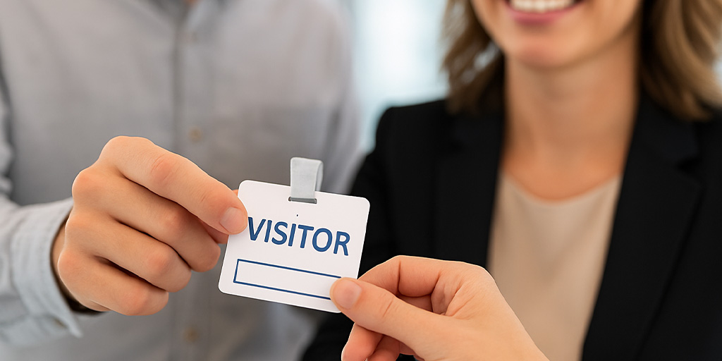 Close-up of a receptionist handing a visitor badge to a professionally dressed guest across a front desk, illustrating a check-in process at a secure facility.